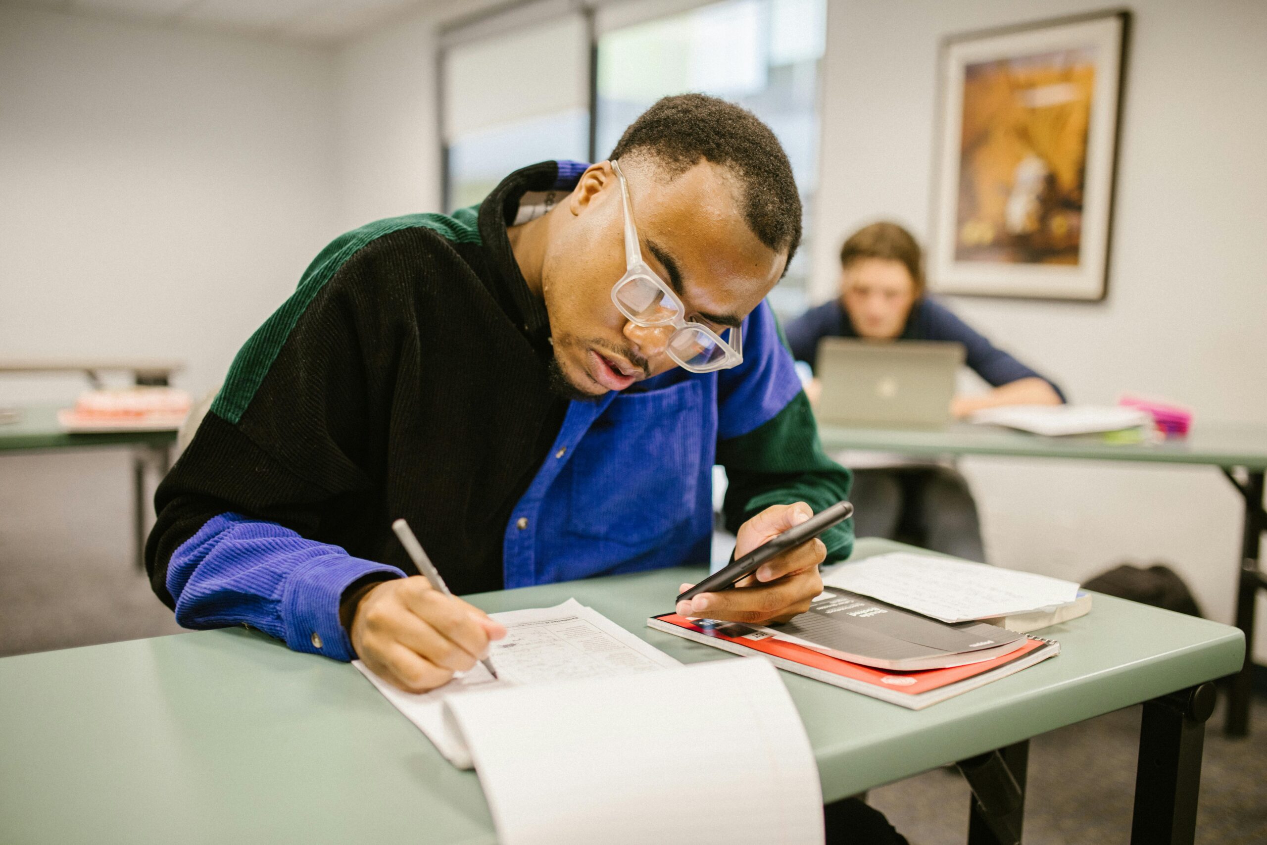 Student sitting at desk using smartphone while preparing for an exam in a classroom setting.