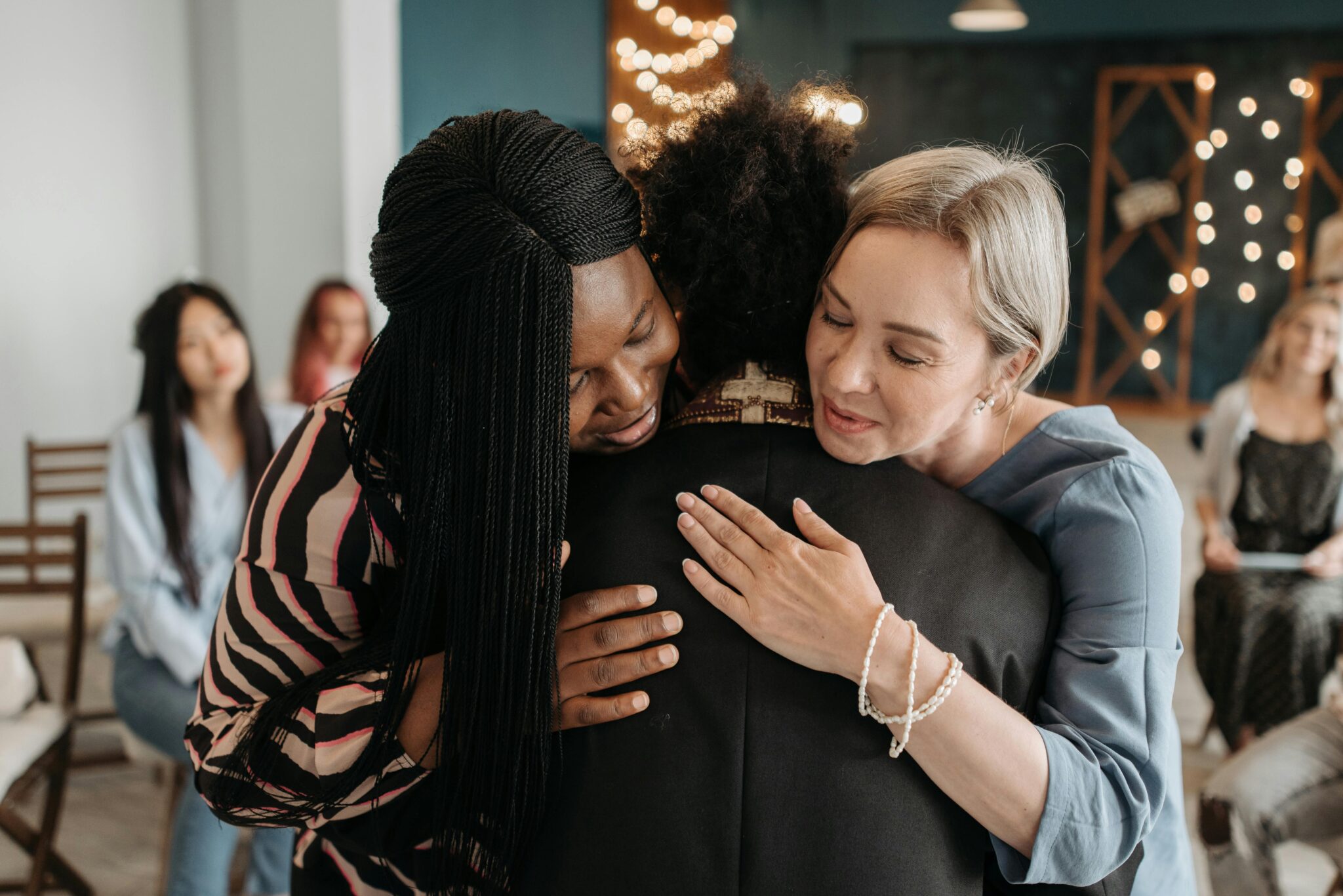 A touching moment as two women embrace a priest during a joyful community gathering indoors.