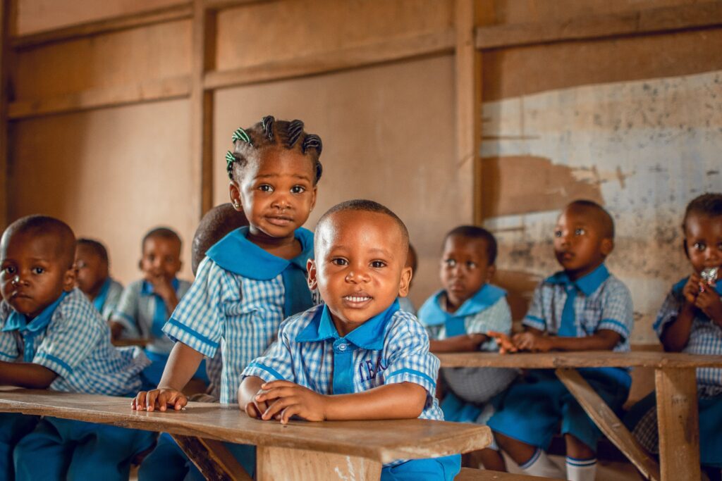 HopeWorks Global Kenya Happy children in a Nigerian classroom wearing uniforms, sitting on wooden benches.