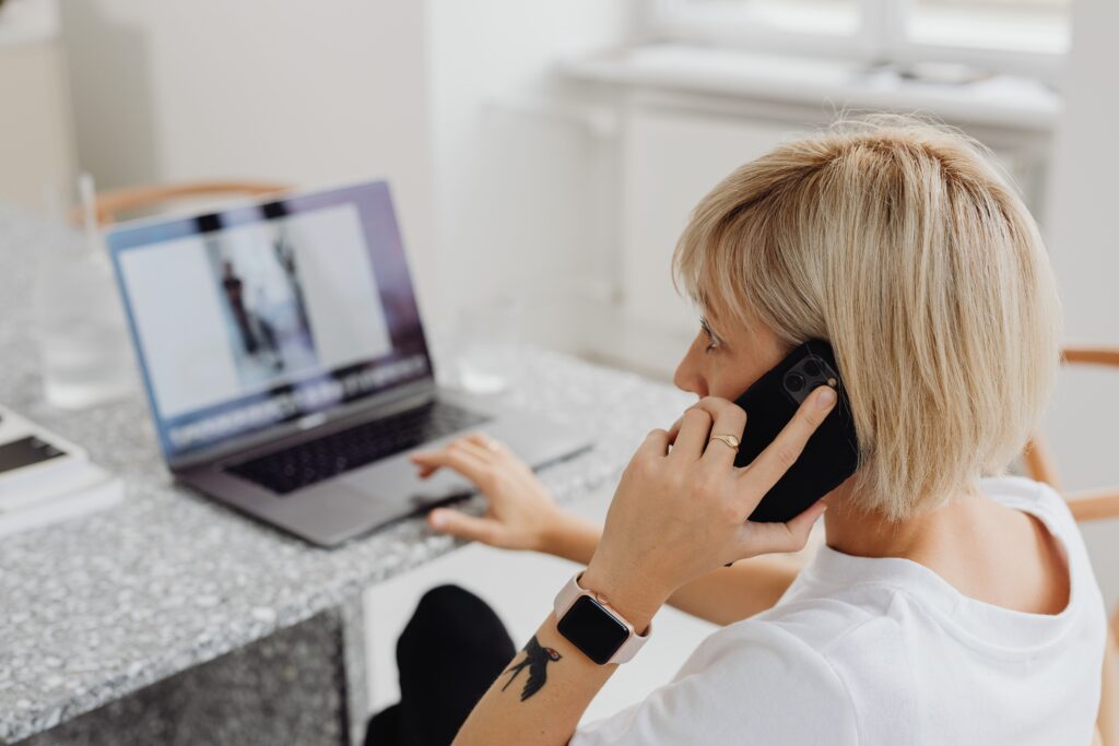 Contact Blonde woman multitasking, working on a laptop and talking on phone indoors.