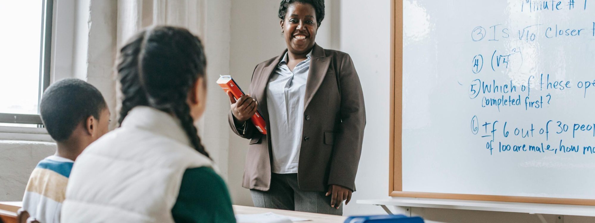 Happy black female teacher standing near whiteboard and explaining lesson to unrecognizable children sitting at wooden table with books and stationery