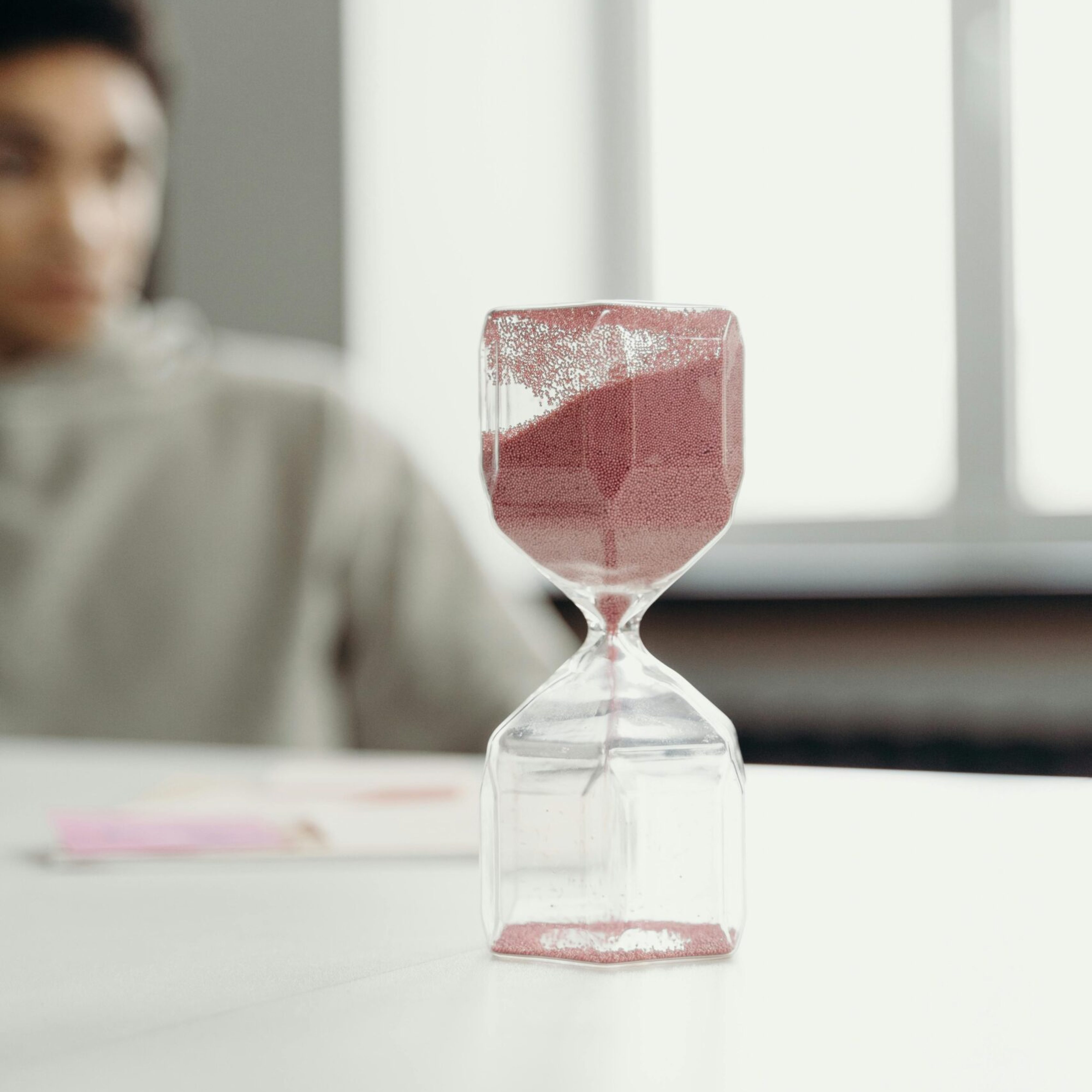 Close-up of an hourglass with red sand against a blurred indoor background, signifying the passage of time.